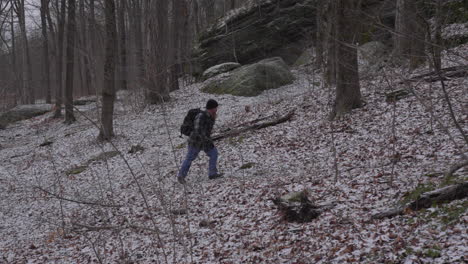 slow-motion-of-a-hiker-with-a-beard-and-wearing-a-flannel-shirt-walks-up-a-woodland-hill-in-early-winter-with-barren-trees-and-a-dusting-of-snow-on-the-ground