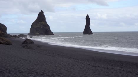 Black-sand-beach-with-towering-sea-stacks-under-a-cloudy-sky-in-Iceland,-waves-lapping-the-shore