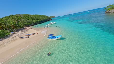 Drone-flying-over-Playa-Ermitano-seashore-and-spectacular-turquoise-sea-water-with-moored-yachts-and-tourists-relaxing-on-beach,-Samana-in-Dominican-Republic
