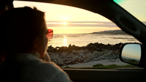 Woman-in-car-points-to-something-in-sea-while-sun-is-setting-over-ocean