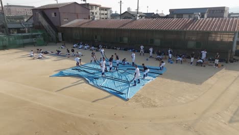 Japanese-baseball-team-trains-with-serpentine-ropes-physical-strength-at-dirt-ground-field-in-school,-Aerial-Drone-View