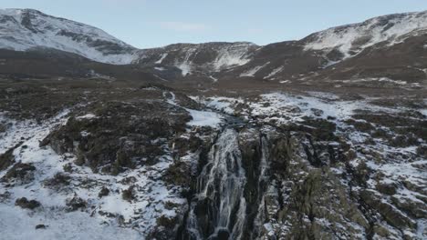 Snow-capped-mountains-and-bride's-veil-waterfall-on-skye-with-cascading-water-and-rugged-terrain,-aerial-view