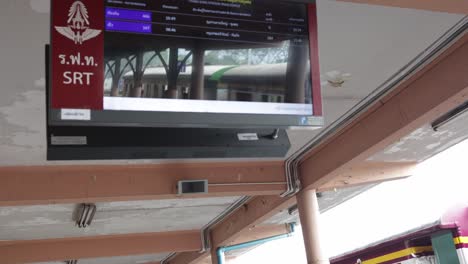 Tilt-down-shot-of-digital-arrival-and-departure-board-showing-timetable-while-a-passenger-train-stopped-at-Thungsong-Junction-Railway-Station