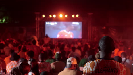 Standing-Man-Wearing-Ivory-Coast-Jersey-Watching-A-Game-With-Fans-In-The-Background,-Africa-Cup-of-Nations-2023,-Abidjan