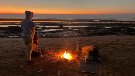 group-of-people-stand-around-the-campfire-seaside-camping-gathering-around-hot-warm-bonfire-sunrise-time-cold-early-morning-in-Qeshm-island-travel-to-marine-adventure-water-sport-surfing-yacht-club