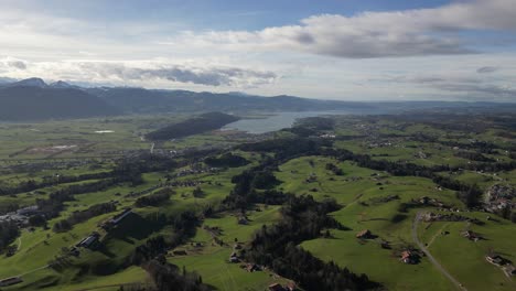 Aufnahme-Eines-Grünen-Offenen-Feldes-Mit-Sporadischen-Dorfhäusern-Im-Sommer-Mit-Blick-Auf-Den-See