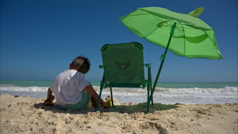 Cámara-Lenta-De-Un-Joven-Latino-Jugando-Solo-En-La-Arena-Junto-A-Su-Silla-De-Playa-Y-Sombrilla-En-Un-Hermoso-Día-Soleado