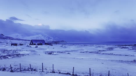 Snow-covered-field-by-the-sea,-with-fence-in-the-foreground,-a-building-in-the-middle-and-mountains-in-the-background
