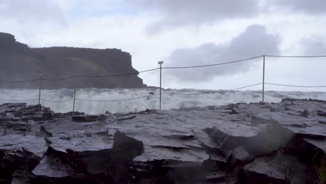 Olas-ásperas-Chocan-Contra-Rocas-Negras-Bajo-Un-Cielo-Nublado,-Acantilado-En-El-Fondo