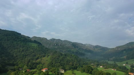 Flug,-Der-Im-Sommer-Eine-Kurve-In-Einem-Tal-Mit-Einem-Dorf-Und-Bauernhäusern-In-Einer-Grünen-Umgebung-Voller-Wiesen-Und-Wälder-Aus-Buchen-Und-Eichen-Mit-Einem-Bewölkten-Himmel-In-Kantabrien,-Spanien-Macht