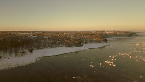 Drone-view-going-towards-the-coast-of-the-Canadian-town-of-Beaumont,-Quebec,-from-the-St-Lawrence-river-Seaway,-on-a-very-cold-Winter-morning
