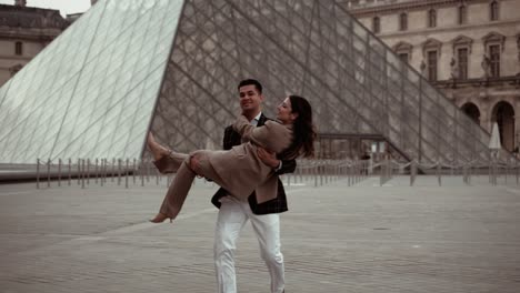 Elegantes-Paar,-Mann-Hält-Seine-Frau-An-Den-Händen,-Während-Er-Sie-An-Der-Pyramide-Des-Louvre-Herumwirbelt,-Umgeben-Von-Den-Barocken-Gebäuden-Der-Königlichen-Residenz-In-Paris,-Frankreich---Moderner-Lebensstil-Und-Anzüge