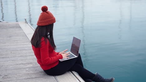 Asian-woman-sits-by-the-water-at-the-port-of-Valencia-ES,-engrossed-in-her-work-on-a-laptop,-her-thoughts-consumed-by-the-task-at-hand,-wearing-a-red-beanie-that-complements-her-red-turtleneck-shirt