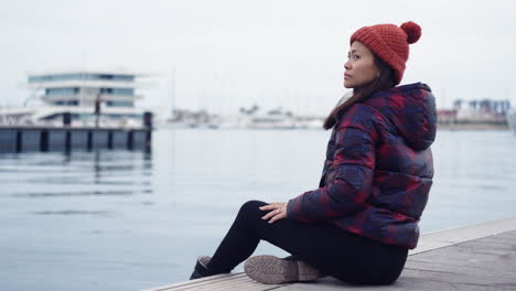 An-Asian-woman-relaxes-by-the-waters-of-Valencia-Port,-Spain,-wearing-a-red-beanie-that-complements-her-matching-checkered-jacket,-casting-a-serene-gaze-towards-the-camera