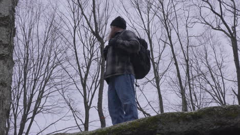 a-man-in-a-flannel-shirt-carrying-a-black-backpack-stands-on-the-apex-of-a-boulder-and-overlooks-the-forest-with-barren-trees-in-the-background-on-a-gray-winter-day