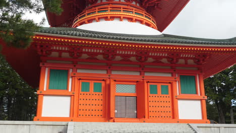 Orange-and-white-Great-Pagoda-in-Koyasan-with-intricate-architectural-details,-surrounded-by-greenery,-under-a-cloudy-sky,-zoom-out