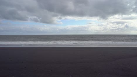 Playa-De-Arena-Negra-Con-Olas-Rompientes-Bajo-Un-Espectacular-Cielo-Nublado-En-Islandia