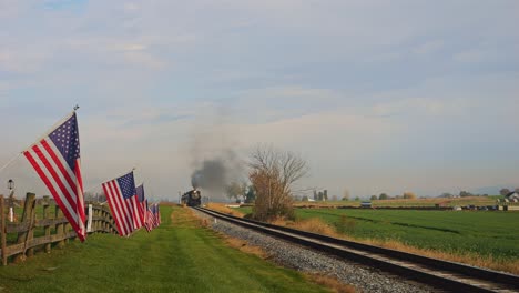 A-View-of-a-Single-Rail-Road-Track,-With-a-Fence-with-America-Flag-on-it,-Gently-Waving-in-the-Wind-as-a-Steam-Train-Approaches,-on-a-Sunny-Autumn-Day