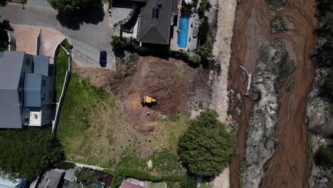 Drone-shot-of-a-yellow-digger-clearing-a-site-in-preparation-for-building-next-to-a-river,-other-houses-and-a-large-tree