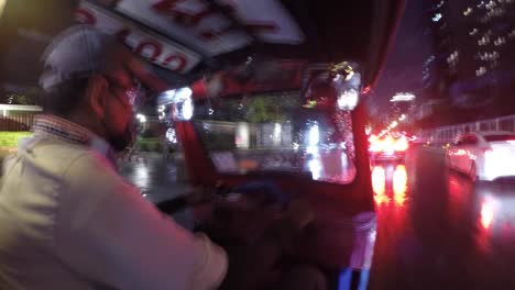 Thailändischer-Tuk-Tuk-Fahrer-Fährt-Auf-Bangkoks-Neonbeleuchteten,-Regenbedeckten-Straßen