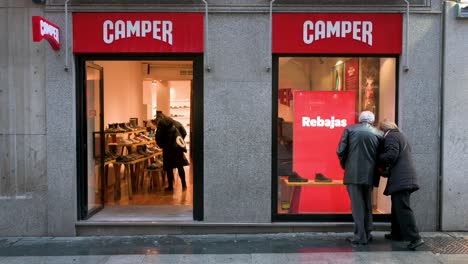 People-window-shopping-at-the-Spanish-multinational-manufacturing-and-footwear-retail-brand-Camper-store-as-pedestrians-walk-past-the-frame-in-Madrid,-Spain