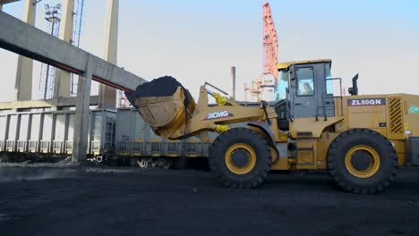 Yellow-wheel-loader-moving-coal-to-an-cargo-train-at-an-industrial-site,-early-morning-light