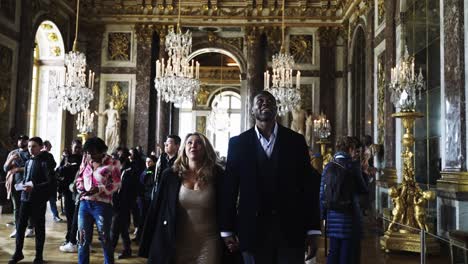 Mixed-race-couple-walking-through-the-famous-Hall-of-Mirros-in-Castle-Versailles-in-Paris-France-with-a-lot-of-chandeliers,-paintings-golden-ornaments-on-the-historical-with-a-lot-of-tourists