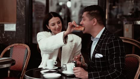 Elegantes-Dunkelhaariges-Paar-Beim-Traditionellen-Französischen-Frühstück,-Während-Die-Frau-Ihren-Mann-Spielerisch-Mit-Einem-Schokoladencroissant-Füttert-Und-Er-In-Einem-Französischen-Café-In-Paris,-Frankreich,-Hineinbeißt