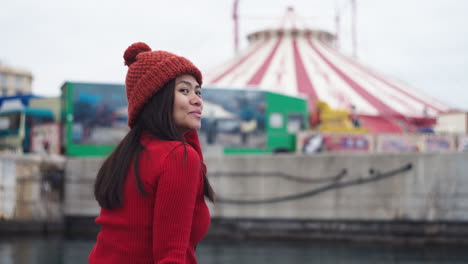 Una-Mujer-Asiática-Sonríe-A-La-Cámara-Mientras-Se-Pone-Un-Gorro-Rojo,-Que-Combina-Perfectamente-Con-Su-Camisa-Roja-De-Cuello-Alto,-En-Un-Puerto-Con-Un-Telón-De-Fondo-De-Temática-Circense-En-El-Puerto-De-Valencia,-España.