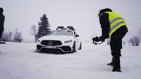 Car-stuck-in-dense-snow-on-race-track,-people-around-watch-and-film-on-camera