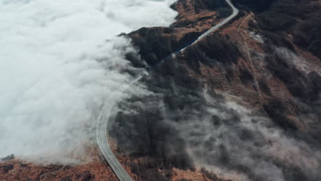 A-winding-road-cutting-through-a-misty-mountain-landscape,-clouds-hovering-close-to-the-ground,-aerial-view