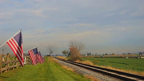 A-View-of-a-Single-Rail-Road-Track,-With-a-Fence-with-America-Flag-on-it,-Gently-Waving-in-the-Wind-as-a-Steam-Train-Approaches,-on-a-Sunny-Autumn-Day