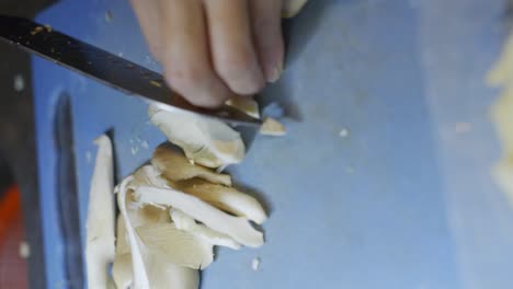 Knife-used-to-cut-mushroom-into-long-slices-on-blue-cutting-board,-filmed-as-vertical-closeup-handheld-slow-motion-style