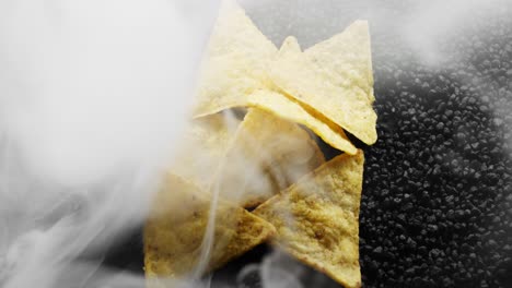 Hand-full-of-tortilla-chips-sitting-on-a-bed-of-black-rocks,-surrounded-by-white-smoke