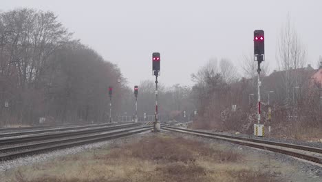 Eisenbahnschienen-Konvergieren-Mit-Roten-Signalen-An-Einem-Nebligen-Tag,-Kahle-Bäume-Im-Hintergrund,-Hauch-Von-Städtischem-Leben