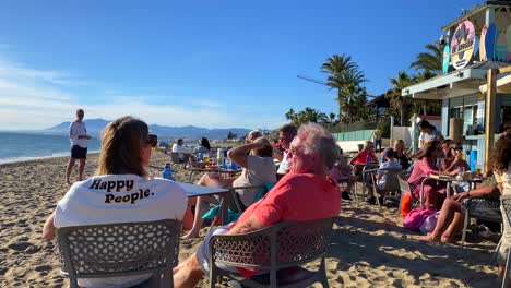 People-sitting,-talking-and-enjoying-life-at-a-beach-restaurant-in-Marbella,-woman-wearing-a-pullover-with-happy-people-words,-sunny-summer-day-in-Spain,-4K-shot