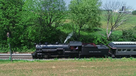 An-Aerial-Side-View-of-a-Steam-Passenger-Train,-Traveling-Thru-Harvested-Farmlands-on-a-Sunny-Day-in-Slow-Motion