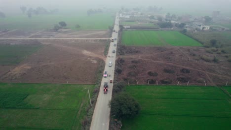 Toma-Aérea-De-Una-Concurrida-Carretera-Rural-En-Alipur,-Pakistán,-Dividiendo-Campos-Verdes,-Con-Tráfico-Y-Neblina.