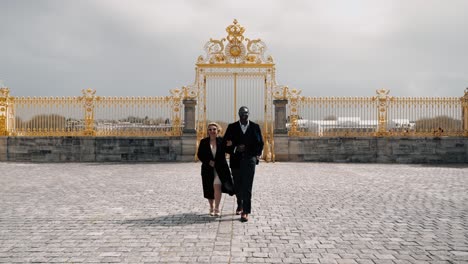 Classy-mixed-race-couple-with-sunglasses-walking-over-the-empty-Cour-d'Honneur---honor-courtyard-of-the-famous-castle-Versailles-in-Paris-France-with-the-golden-honour-gate-in-the-background---wide