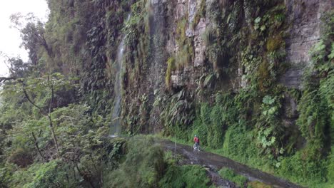 Luftaufnahme-Folgt-Radfahrer,-Wo-Ein-Wasserfall-Auf-Die-Yungas-Road-Stürzt,-Bolivien