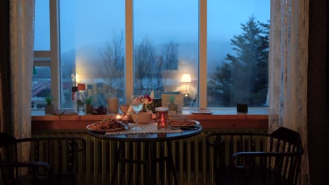 Cozy-evening-dinner-for-two-by-window-with-mood-lighting,-pasta-and-drinks-on-table,-overcast-outdoor-view