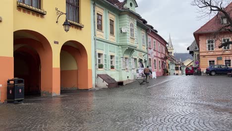 Old-man-carrying-a-big-wood-log-during-rainy-day-through-historical-city-Sighisoara-,-Romania