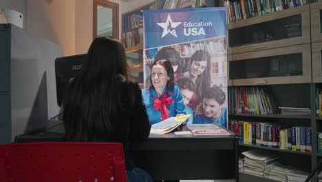 Moving-shot-of-a-Caucasian-woman-in-uniform-sitting-at-a-desk-giving-information-to-another-woman-in-an-office-with-books-and-a-sign-that-says-"Education-USA