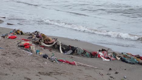 Piles-of-discarded-clothes-and-fabric,-fast-fashion,-washed-ashore-onto-sandy-beach-from-ocean-with-gentle-waves-on-a-tropical-island-destination