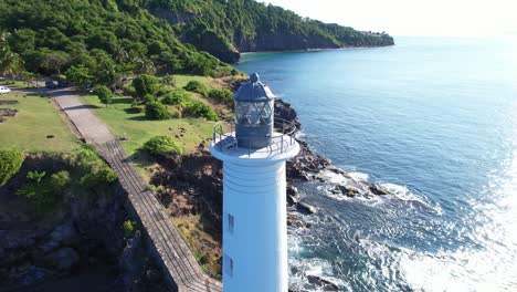 Drohne-Fliegt-Um-Die-Spitze-Des-Leuchtturms-Am-Vieux-Fort-Mit-Zerklüfteter-Küste-Im-Hintergrund,-Guadeloupe