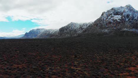 Aerial-drone-shot-along-the-desert-with-snowcapped-mountains,-clouds-and-blue-skies-in-the-background