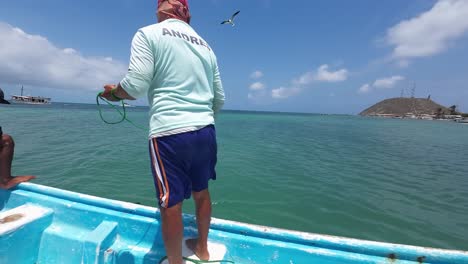 Fisherman-casting-net-from-boat-on-sunny-day,-with-clear-blue-water-and-a-ship-in-the-distance,-dynamic-action-shot