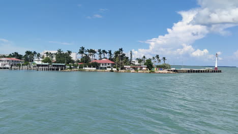 Una-Vista-Aérea-Revela-Una-Isla-Adornada-Con-Un-Faro-Y-Un-Vibrante-Astillero,-Con-El-Telón-De-Fondo-Del-Exuberante-Paisaje-Caribeño,-Donde-Los-Cocoteros-Y-Las-Palmeras-Se-Mecen-En-Armonía.