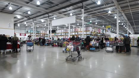Busy-Costco-checkout-lines-with-shoppers-and-their-trolleys,-fluorescent-lighting,-indoors