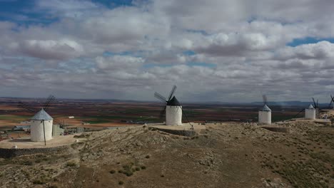 lateral-flight-with-drone-visualizing-four-old-windmills-visited-by-people-doing-tourism-with-a-background-of-farmland-and-population,-on-a-cloudy-spring-day-in-Toledo-Spain